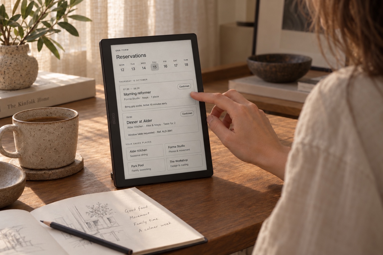 A person using a small e-ink display with a monochrome reservations view at a warm walnut writing desk.